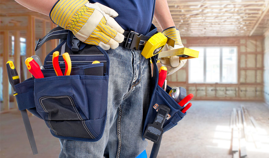 Man with tool belt A construction worker wearing gloves and a tool belt filled with various tools, holding a measuring tape in a partially built house with exposed wooden framing and bright natural light.