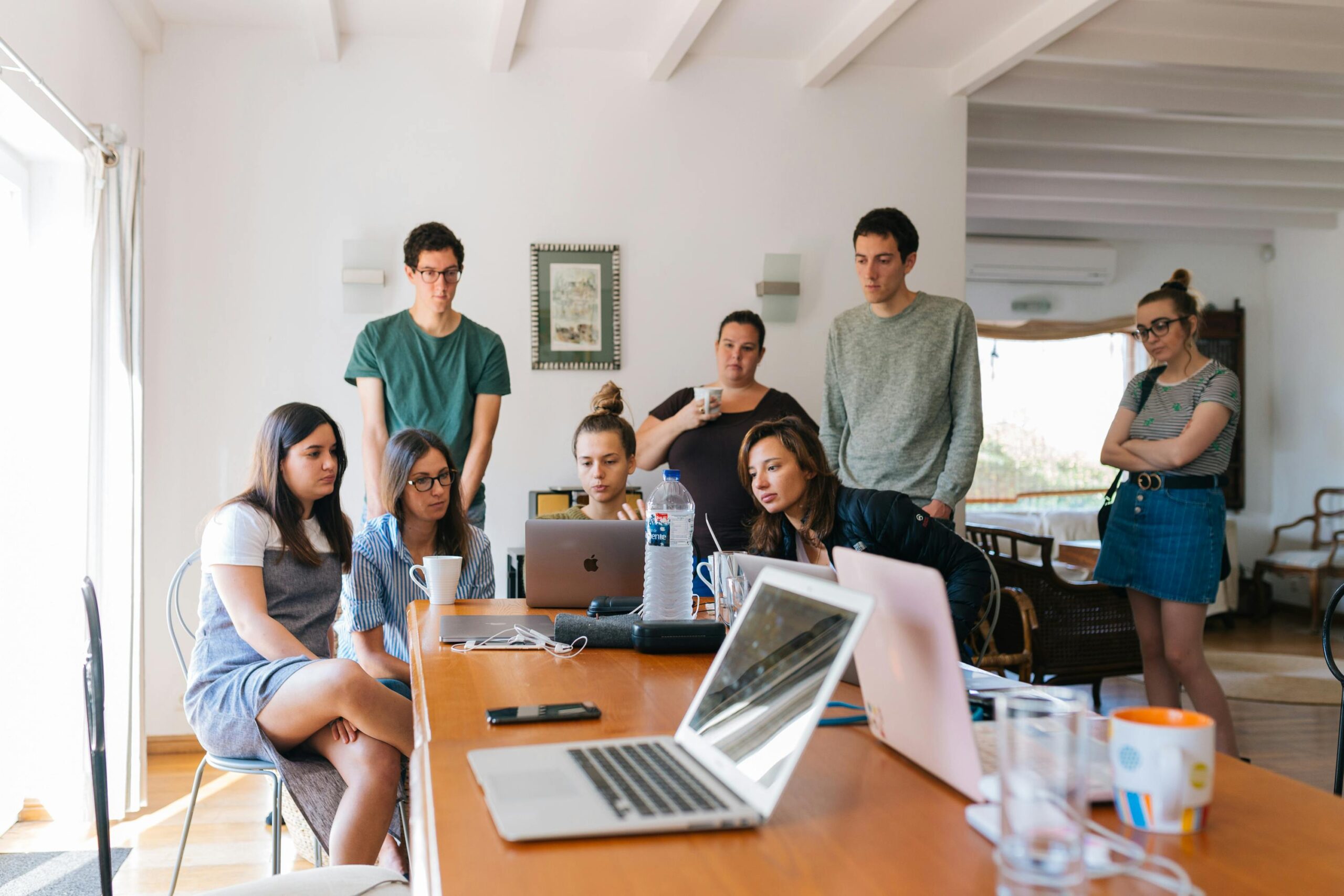 Eight people from the teamwin managed security & IT services team are gathered around a table with laptops and coffee cups, focused on a screen in a bright, modern room.