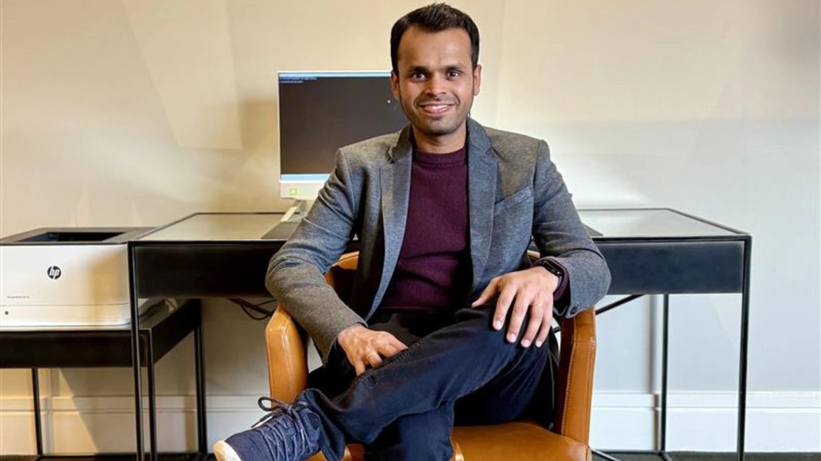 Naman (1) A man in a blazer and maroon sweater sits on a brown chair, smiling at the camera. Behind him are a desk, a computer monitor, and a printer against a light-colored wall.