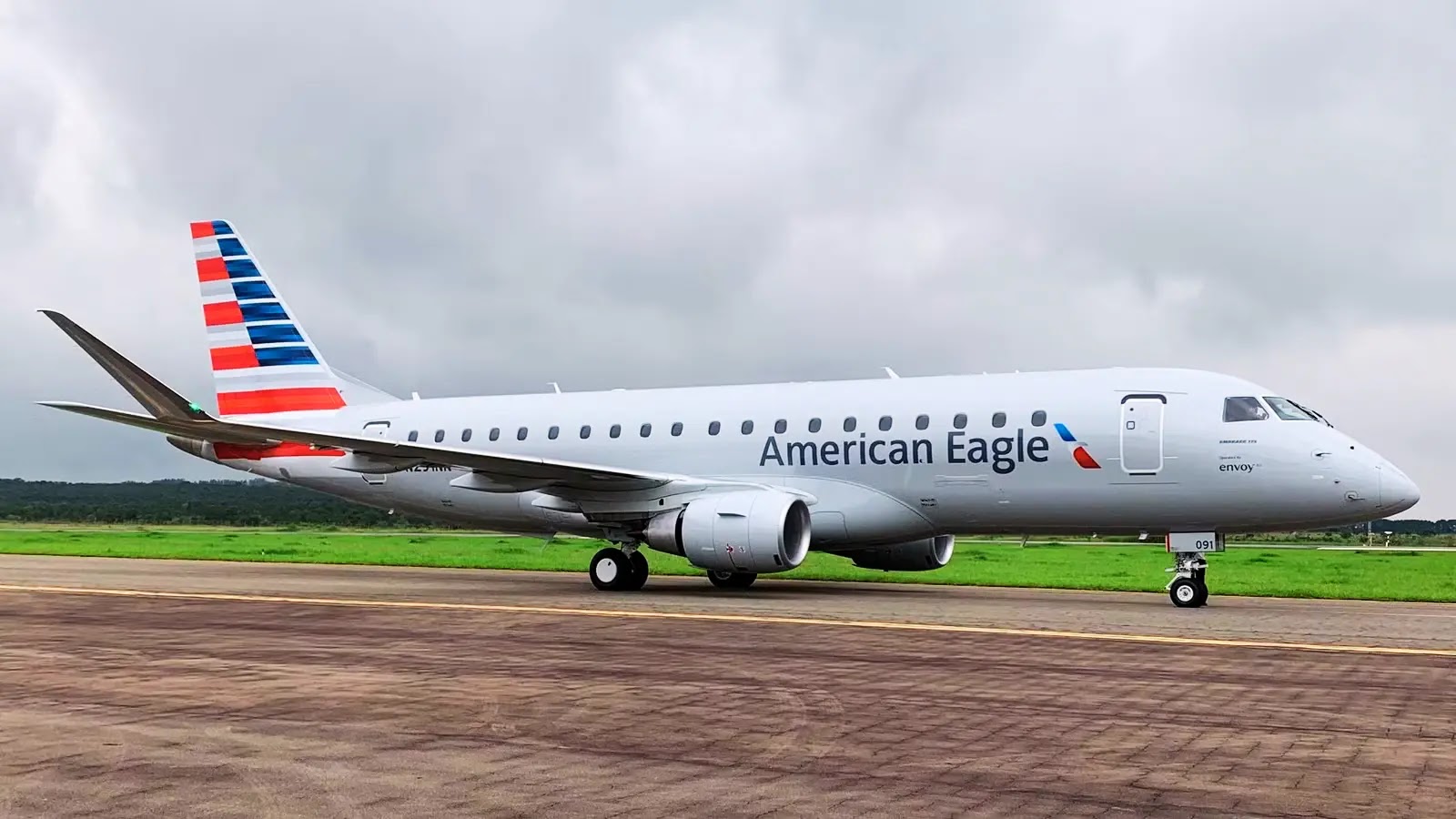 Envoy Compromised Oracle Campaign A white American Eagle passenger jet with red, white, and blue stripes on the tail is parked on an airport runway with green grass and cloudy skies in the background.