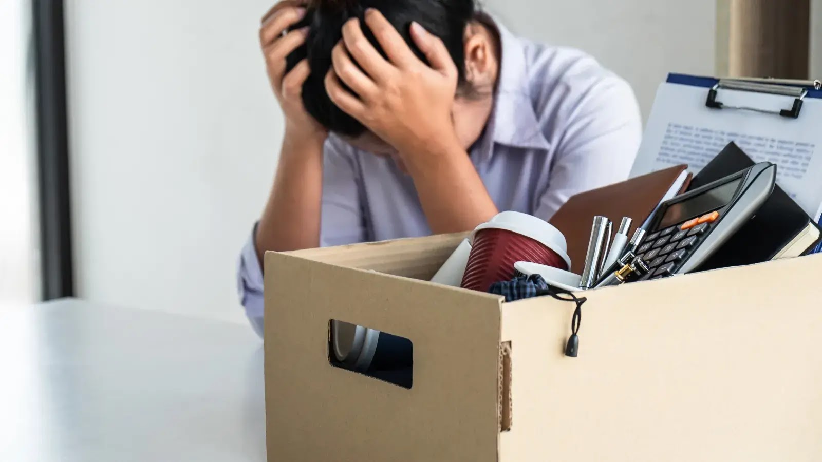A person sits at a desk with their head in their hands, looking distressed. In the foreground is a cardboard box filled with office supplies, suggesting job loss or resignation.
