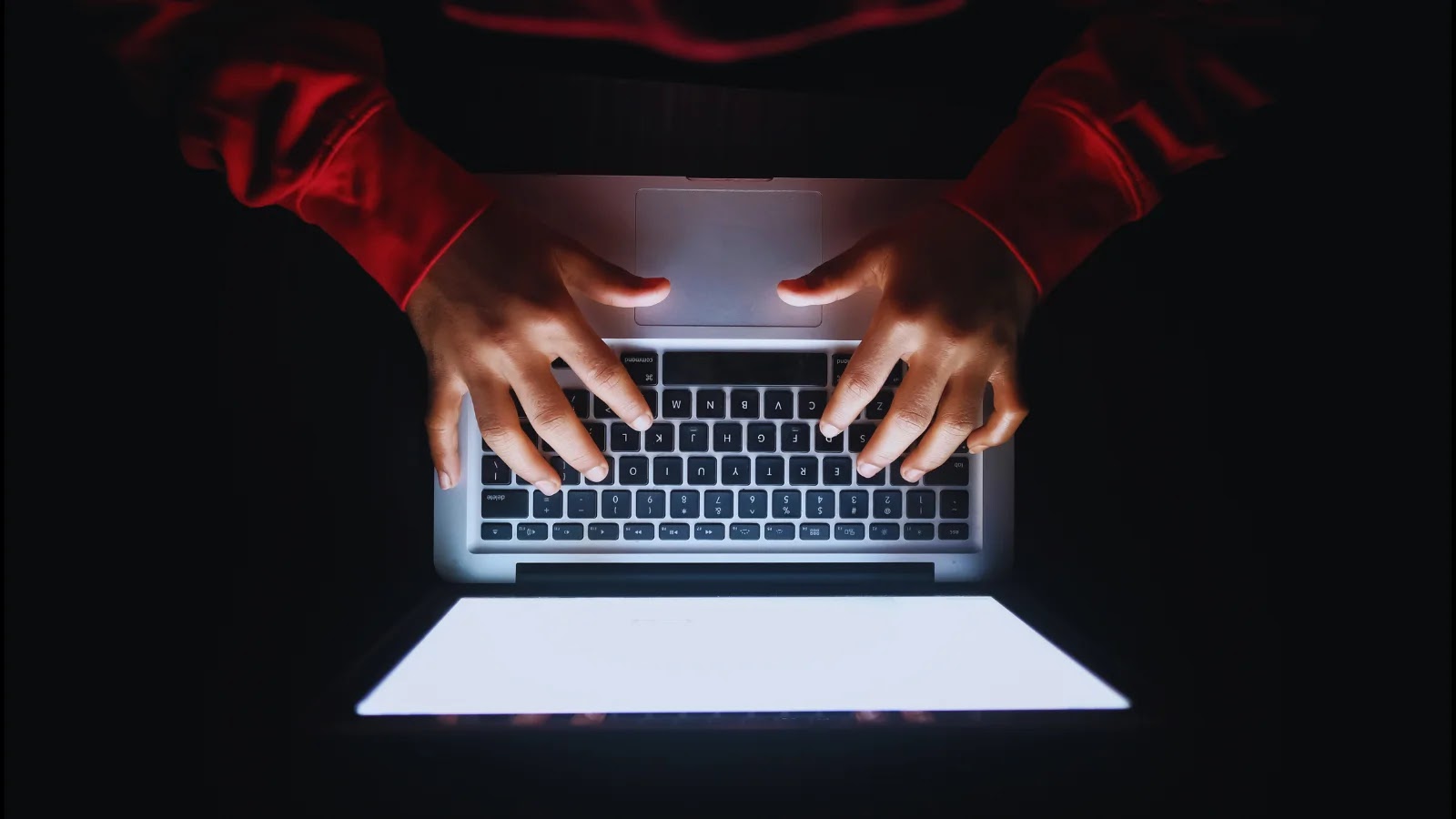 First Large-scale Cyberattack Using AI A person wearing a red long-sleeve shirt types on a laptop keyboard in a dark environment, illuminated only by the bright screen.