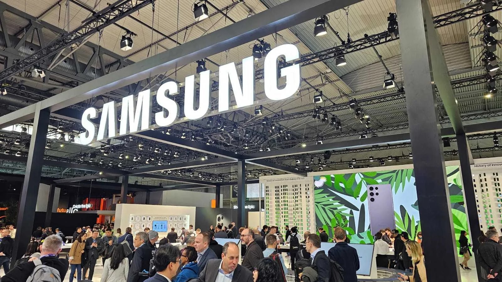 Spyware on Samsung Devices A busy Samsung exhibition booth at a tech event, with a large illuminated Samsung sign overhead, crowds of people, and digital displays showing smartphones and colorful graphics.