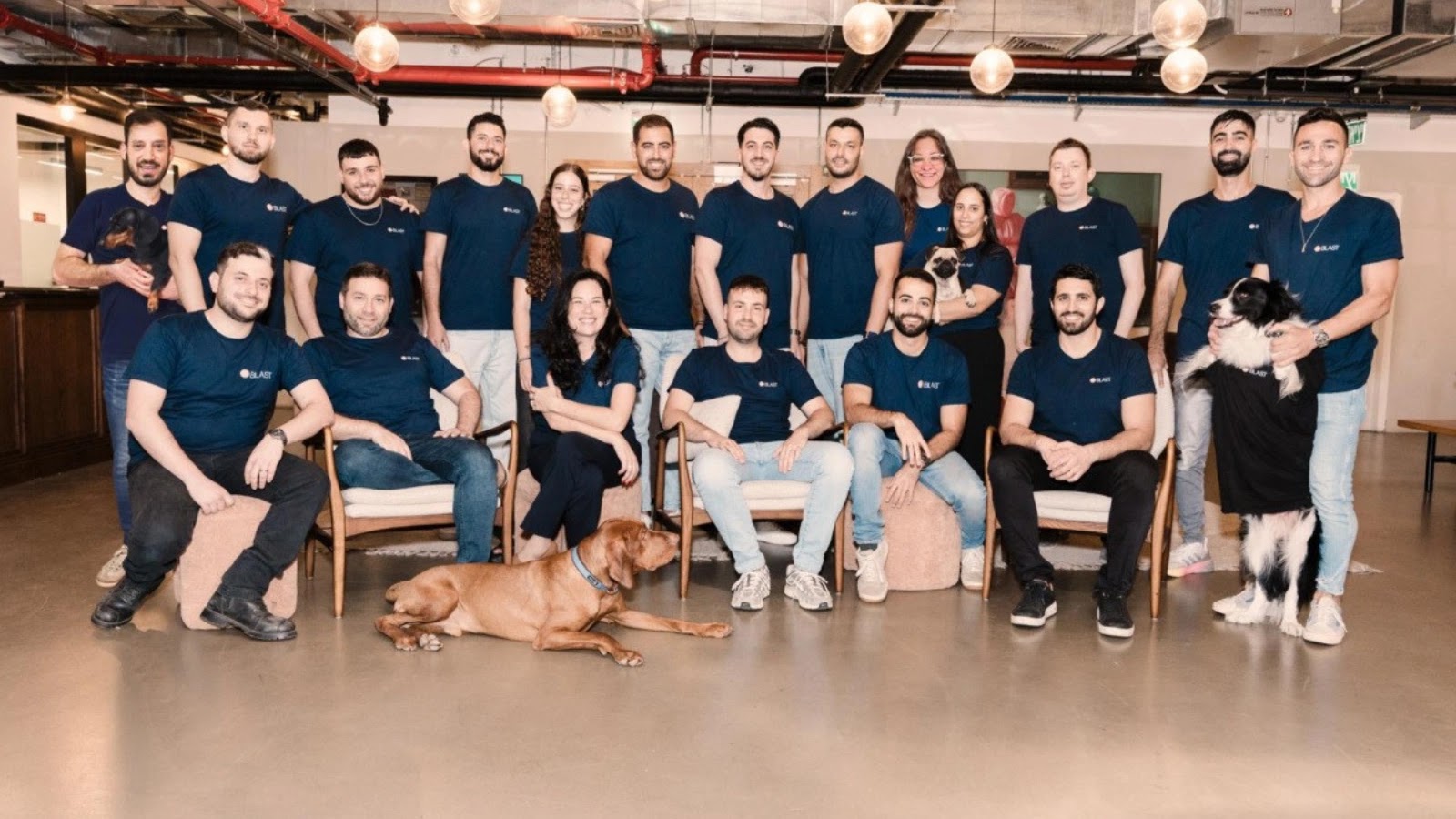 A group of 21 people wearing matching navy blue shirts pose for a photo in an office space. They are smiling, and two dogs are also present—one lying on the floor, the other sitting upright wearing a black shirt.
