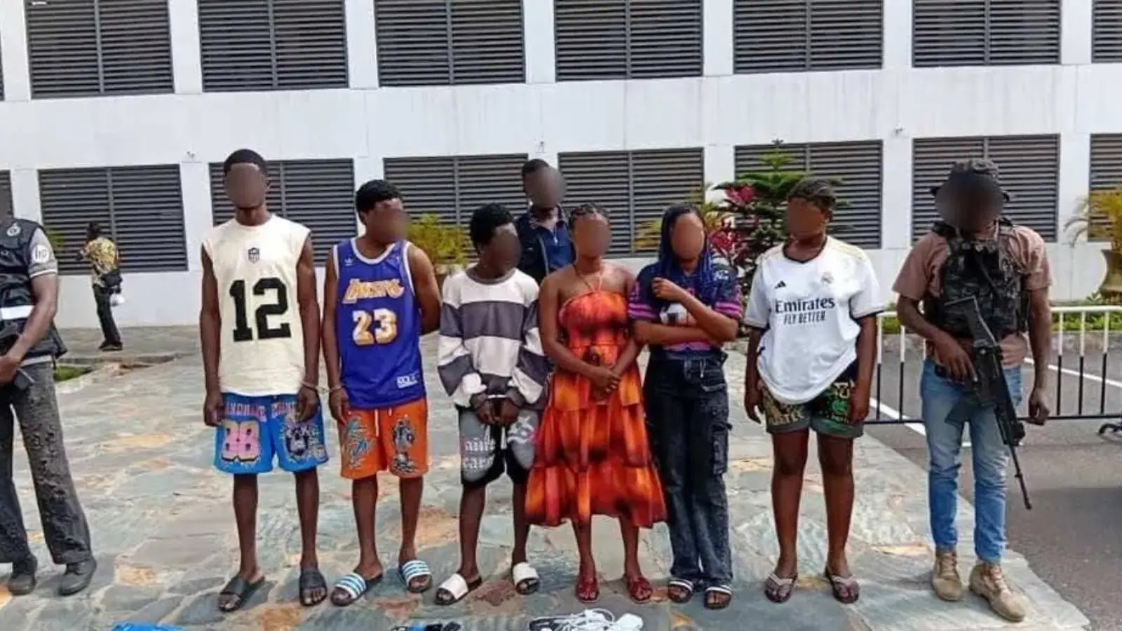 A group of seven people stand in a line outside a building; their faces are blurred. Some wear casual clothes, one wears a dress, and two individuals in uniform stand on either end, appearing to be security or police officers.