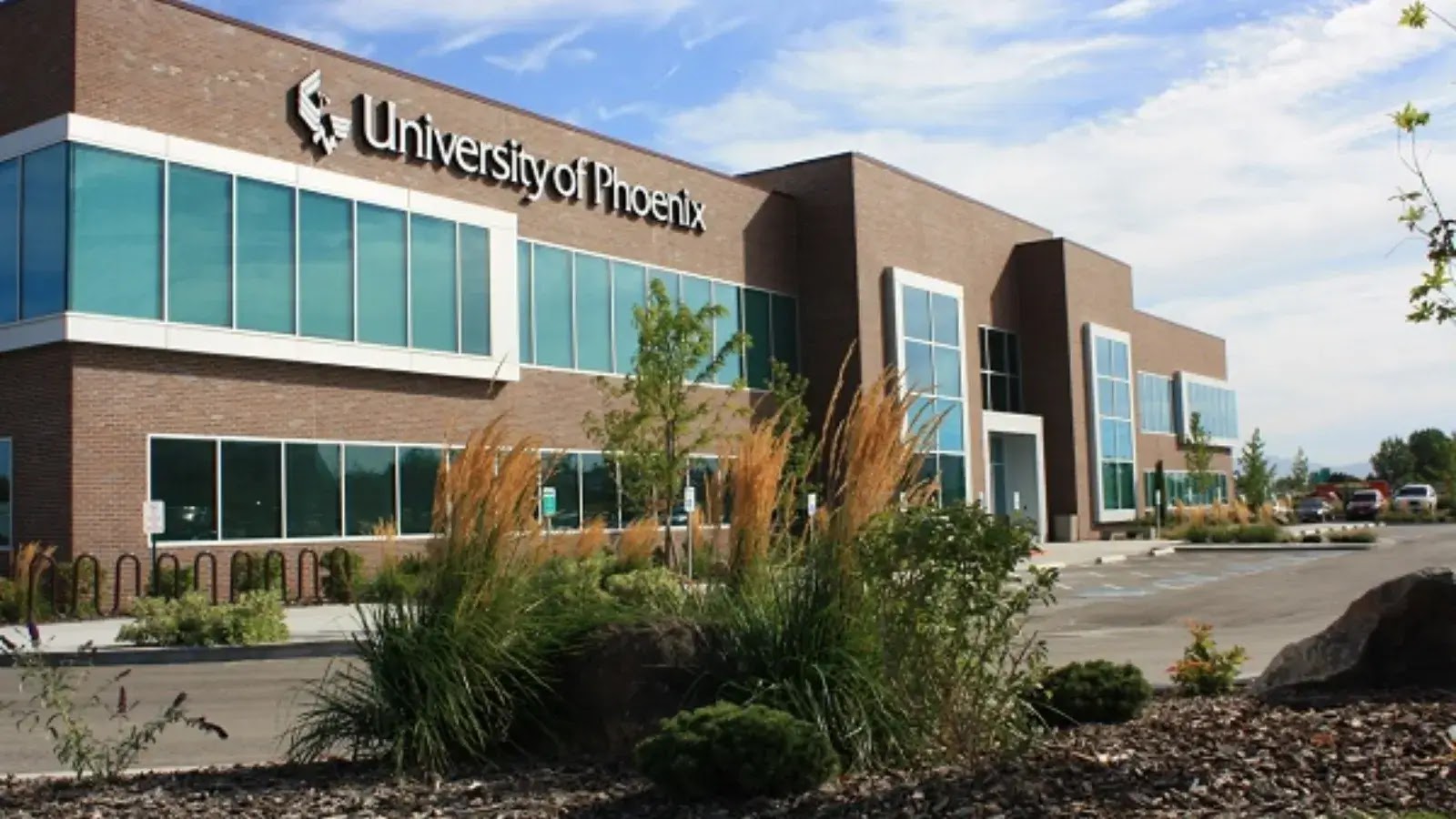A modern two-story brick building with large windows and a sign reading University of Phoenix on the facade, surrounded by landscaping and a mostly empty parking lot under a blue sky.