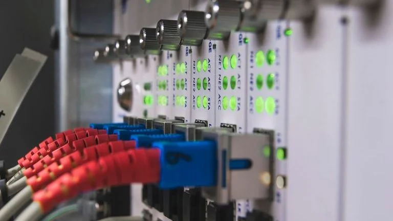 Close-up of network server equipment with glowing green LED lights and multiple connected cables, including red and blue fiber optic cables plugged into ports.