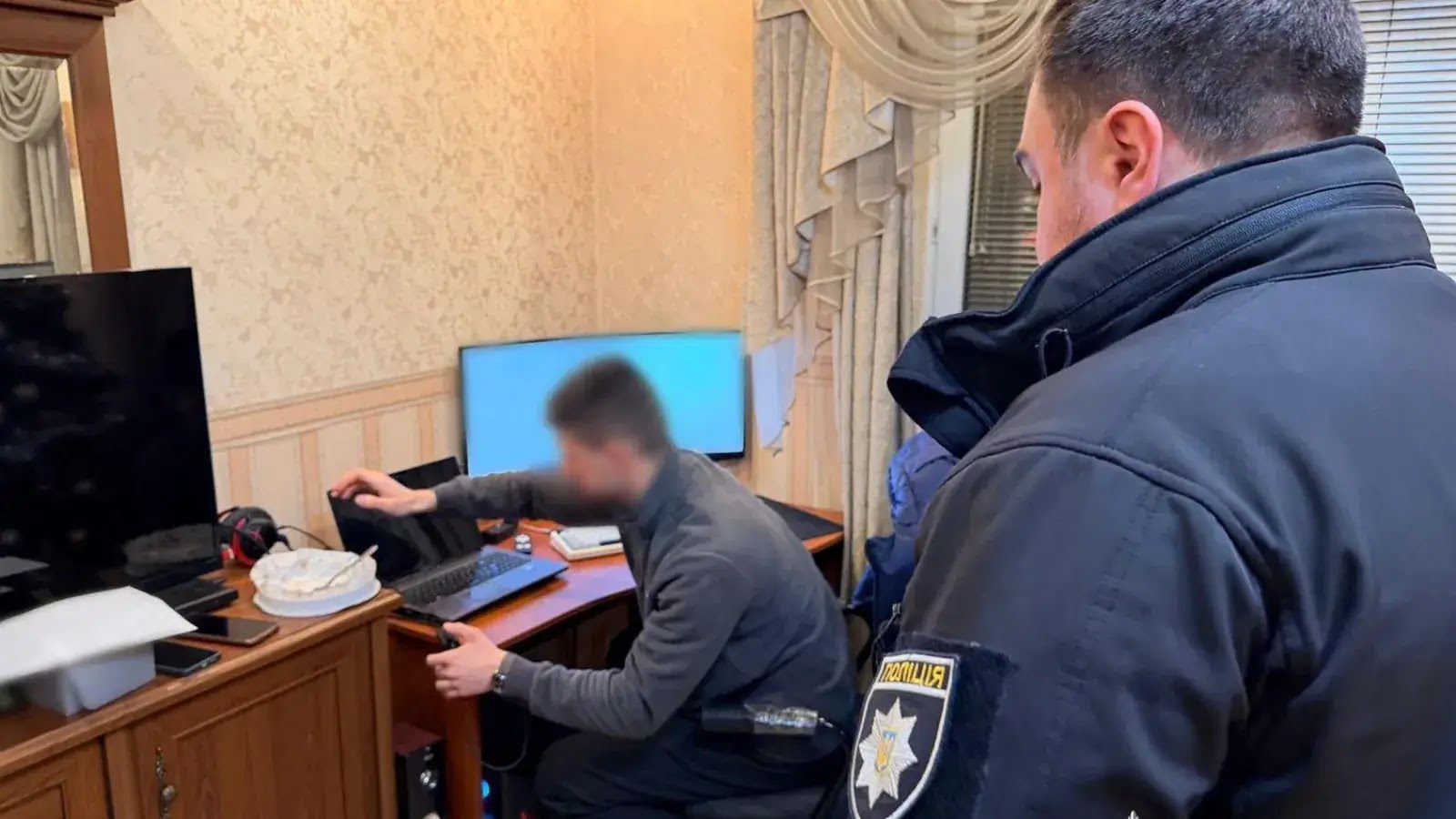 A police officer stands next to a man at a desk with electronic devices and documents; the mans face is blurred, and the room appears to be an office or study.