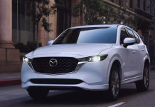 A white Mazda CX-5 SUV is driving on a city street with buildings and trees in the background, headlights on, during the day.