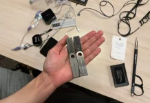 A hand holds two rectangular metal objects with holes near one end. On the table are various items, including tangled wires, earphones, and small black cases.