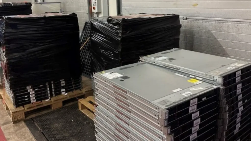 Stacks of servers on pallets, some wrapped in black plastic, in a warehouse.