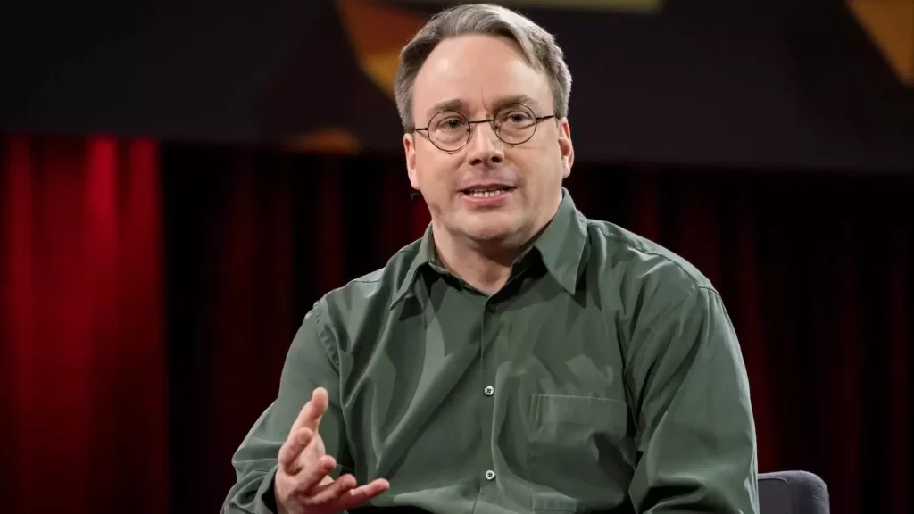 Linus-Torvalds-says-AI-Bug-reports-have-made-Linux-security-mailing-list-unmanageable–1024×576 A man with short hair and glasses, wearing a green button-up shirt, speaks while seated on stage with a red and black background.