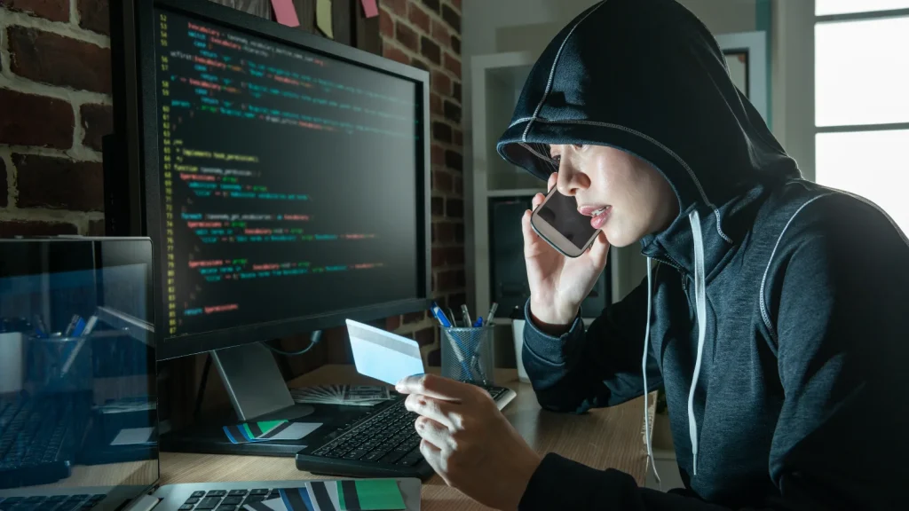 U.S.-Executives-Plead-Guilty-1024×576 A person in a hoodie is sitting at a desk, holding a credit card and talking on the phone, with a computer screen displaying code in front of them.