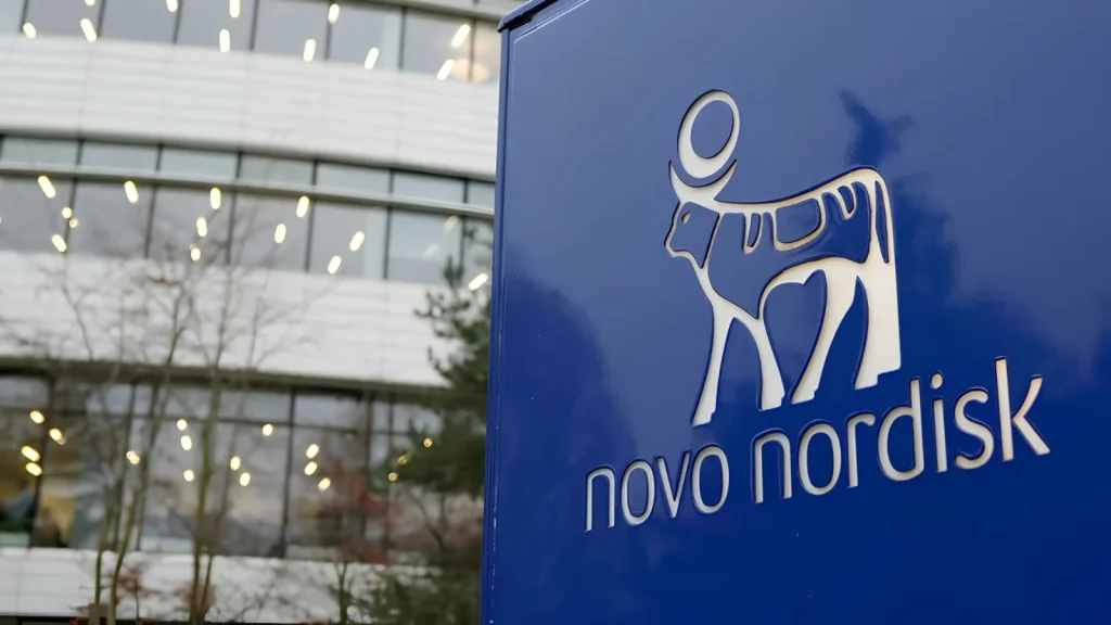 Novo-Nordisk-Confirms-Cyber-Attack-1024×576 A blue sign with the Novo Nordisk logo—an abstract bull and text—stands in front of a modern building with large windows and illuminated lights. Trees are visible in the background.