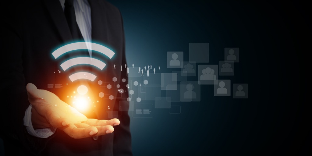 Wi-Fi security for BYOD A person in a suit holds out their hand with a glowing Wi-Fi symbol above it, representing connectivity, technology, and digital networking, against a dark background with faint icons.